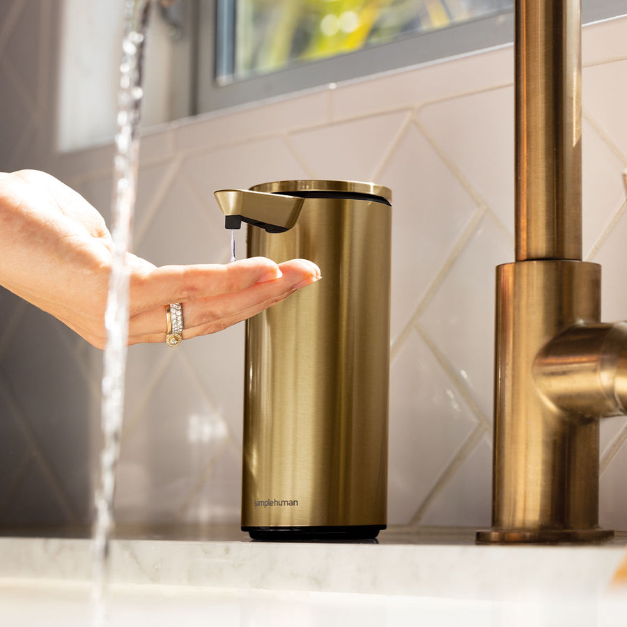 Simplehuman Brushed Brass Sensor Pump dispensing soap onto a hand by a faucet, touch-free operation.